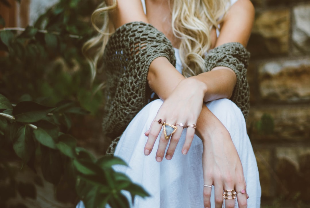 Une femme aux longs cheveux blonds est assise à l'extérieur, vêtue d'une robe blanche de luxe et d'un châle en crochet, avec plusieurs bagues aux doigts.