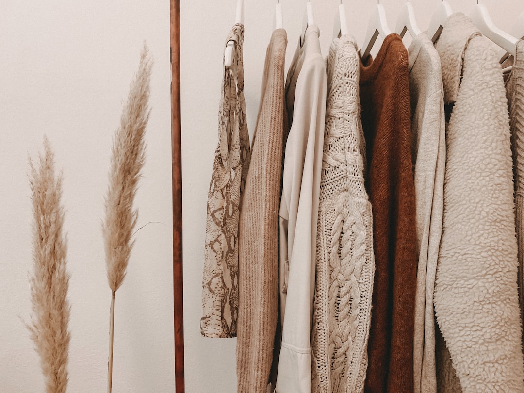 Une rangée de pulls et de vestes de luxe aux couleurs neutres est suspendue à des cintres blancs à côté de deux tiges d'herbe de la pampa séchée.